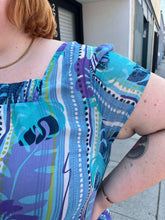 Load image into Gallery viewer, Close up view of the cap sleeves and square neckline of a size 22 Avenue vintage aqua blue, purple, green, and white tropical pattern cap sleeve blouse on a size 22/24 model. The photo is taken outside in natural lighting.
