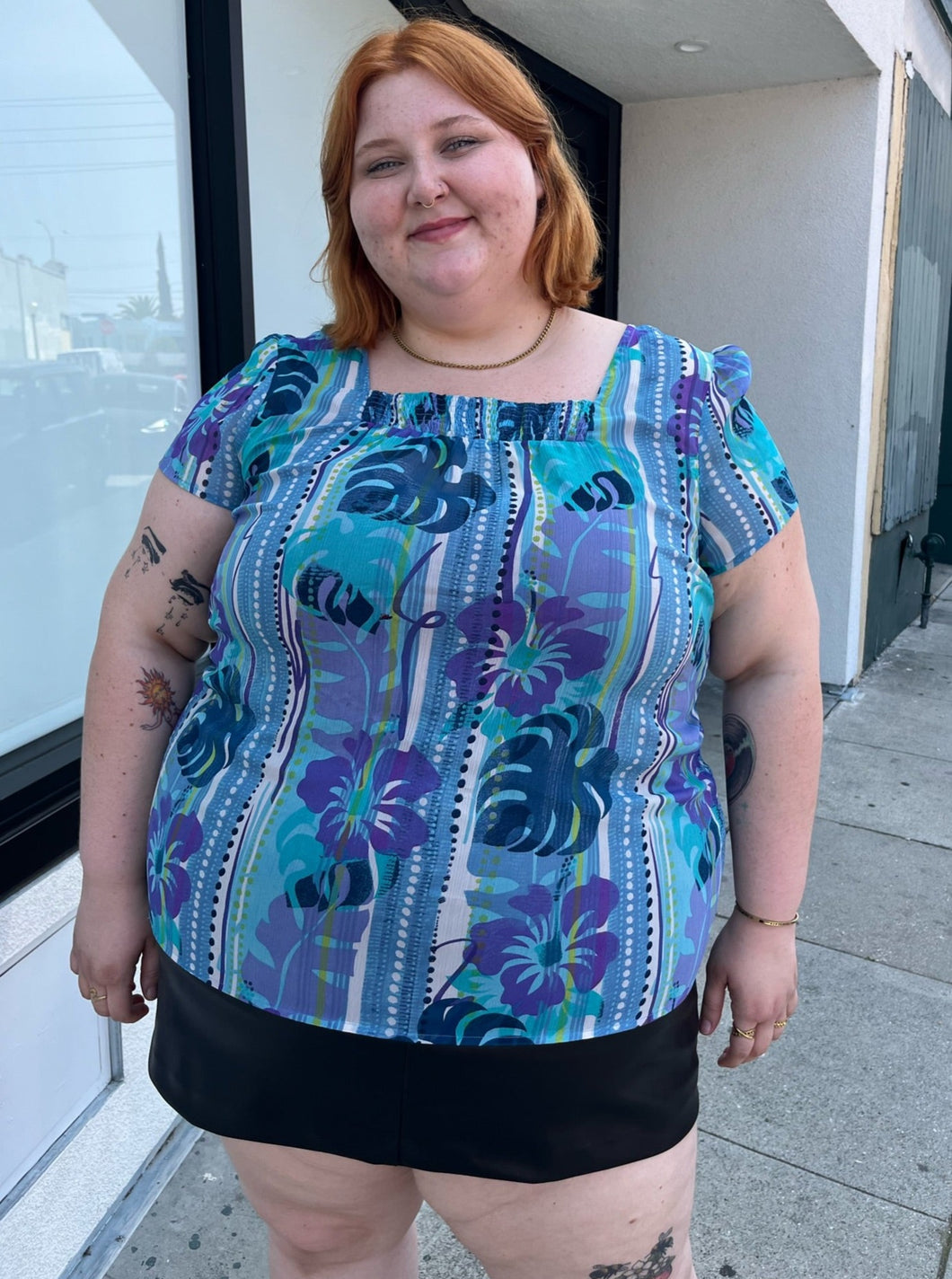 Front view of a size 22 Avenue vintage aqua blue, purple, green, and white tropical pattern cap sleeve blouse styled over a black mini skirt on a size 22/24 model. The photo is taken outside in natural lighting.