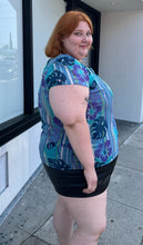 Load image into Gallery viewer, Side view of a size 22 Avenue vintage aqua blue, purple, green, and white tropical pattern cap sleeve blouse styled over a black mini skirt on a size 22/24 model. The photo is taken outside in natural lighting.
