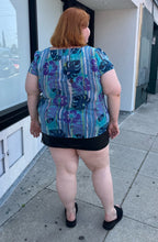 Load image into Gallery viewer, Full-body back view of a size 22 Avenue vintage aqua blue, purple, green, and white tropical pattern cap sleeve blouse styled over a black mini skirt with black slides on a size 22/24 model. The photo is taken outside in natural lighting.
