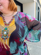 Load image into Gallery viewer, Close up view of the abstract pattern and sheer-lean of the fabric of a size 20-24 purple, yellow, blue, pink, and black abstract pattern 100% silk blouse with beaded and bejeweled neckline on a size 22/24 model. The photo is taken outside in natural lighting.
