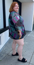 Load image into Gallery viewer, Full-body side view of a size 20-24 purple, yellow, blue, pink, and black abstract pattern 100% silk blouse with beaded and bejeweled neckline styled with a black mini skirt and black slides on a size 22/24 model. The photo is taken outside in natural lighting.
