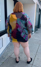 Load image into Gallery viewer, Full-body back view of a size 20-24 purple, yellow, blue, pink, and black abstract pattern 100% silk blouse with beaded and bejeweled neckline styled with a black mini skirt and black slides on a size 22/24 model. The photo is taken outside in natural lighting.
