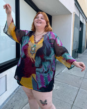 Load image into Gallery viewer, Front view of a size 20-24 purple, yellow, blue, pink, and black abstract pattern 100% silk blouse with beaded and bejeweled neckline on a size 22/24 model. The photo is taken outside in natural lighting.
