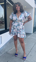 Load image into Gallery viewer, Full-body front view of a size 3X Fashion Nova light blue, white, and purple floral wrap mini dress with matching belt and puff sleeves styled with blue heels on a size 16/18 model.
