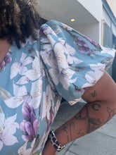 Load image into Gallery viewer, Close up view of the puff sleeve detail of a size 3X Fashion Nova light blue, white, and purple floral wrap mini dress with matching belt and puff sleeves on a size 16/18 model.
