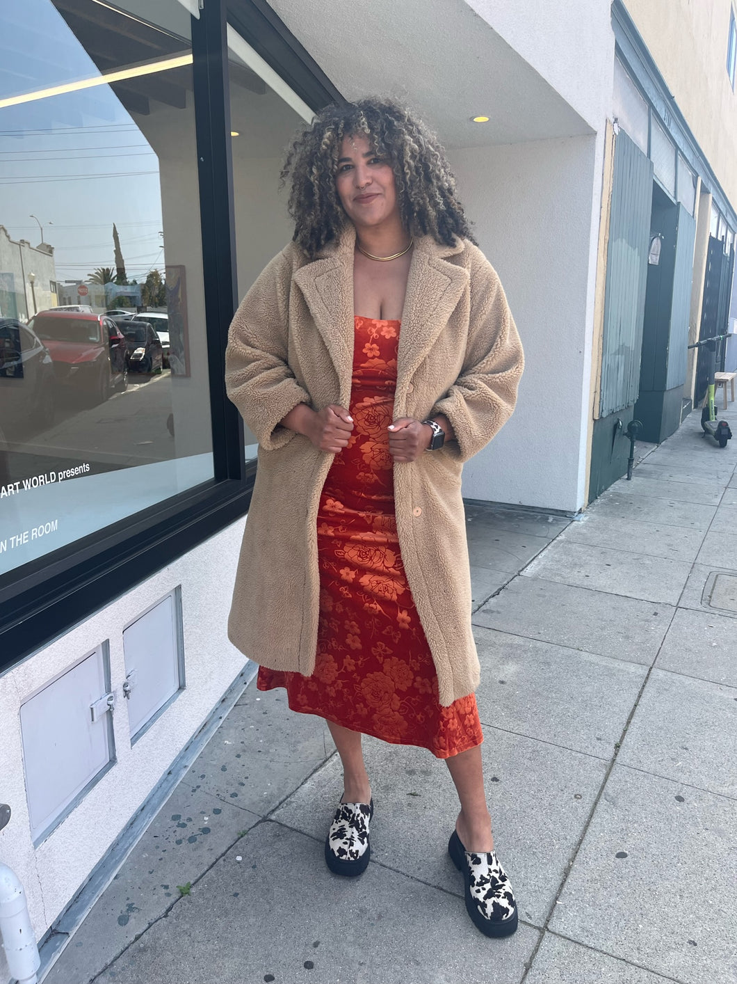 Full-body front view of a size  1X Stand Studio Teddy Bear Jacket on a size 16/18 model. They are wearing a rust orange floral dress beneath and cow print clog shoes. The model is holding the lapels of the jacket. 