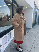 Load image into Gallery viewer, A side view of a size 1X Stand Studio Teddy Bear Jacket on a size 16/18 model. They are wearing a rust orange floral dress beneath and cow print clog shoes. The model has a hand in the pocket of the jacket. 
