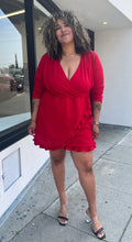 Load image into Gallery viewer, Full-body front view of a size 22 Pretty Little Thing red wrap mini dress with ruffles and tulip hem styled with the sleeves pushed up and black heels on a size 16/18 model. The photo is taken outside in natural lighting.
