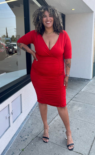Full-body front view of a size 3X Poseshe vibrant red ruched v-neck bodycon midi dress with three-quarter length sleeves styled with black and clear heels on a size 16/18 model. The photo is taken outside in natural lighting.