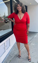 Load image into Gallery viewer, Additional full-body front view of a size 3X Poseshe vibrant red ruched v-neck bodycon midi dress with three-quarter length sleeves styled with black and clear heels on a size 16/18 model. The photo is taken outside in natural lighting.
