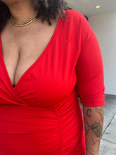Load image into Gallery viewer, Close view of the v-neck, tummy ruching, and sleeve length of a size 3X Poseshe vibrant red ruched v-neck bodycon midi dress with three-quarter length sleeves on a size 16/18 model. The photo is taken outside in natural lighting.
