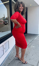 Load image into Gallery viewer, Full-body side view of a size 3X Poseshe vibrant red ruched v-neck bodycon midi dress with three-quarter length sleeves styled with black and clear heels on a size 16/18 model. The photo is taken outside in natural lighting.
