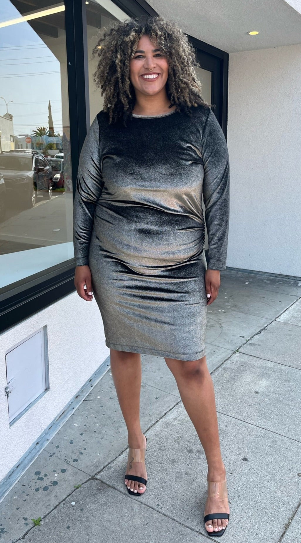 Full-body front view of a size 2X Rachel Roy metallic brown-gold velvet long sleeve bodycon midi dress with ruching at the sides styled with black and clear heels on a size 16/18 model. The photo is taken outside in natural lighting.