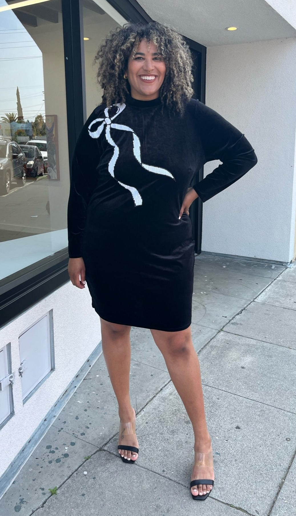 Full-body front view of a size 24 Eloquii black velour long sleeve midi dress with a white sequin bow graphic at the shoulder styled with black heels on a size 16/18 model. The photo is taken outside in natural lighting.