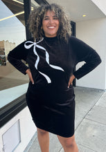 Load image into Gallery viewer, Front view of a size 24 Eloquii black velour long sleeve midi dress with a white sequin bow graphic at the shoulder on a size 16/18 model. The photo is taken outside in natural lighting.
