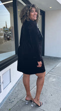 Load image into Gallery viewer, Full-body side view of a size 24 Eloquii black velour long sleeve midi dress with a white sequin bow graphic at the shoulder styled with black heels on a size 16/18 model. The photo is taken outside in natural lighting.
