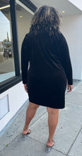 Load image into Gallery viewer, Full-body back view of a size 24 Eloquii black velour long sleeve midi dress with a white sequin bow graphic at the shoulder styled with black heels on a size 16/18 model. The photo is taken outside in natural lighting.
