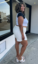 Load image into Gallery viewer, Full-body side view of a pair of size 22 Eloquii lightly distressed cream colored denim shorts styled with a white crop and white heels on a size 16/18 model. The photo is taken outside in natural lighting.
