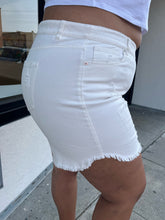 Load image into Gallery viewer, Side view of a pair of size 22 Eloquii lightly distressed cream colored denim shorts styled with a white crop and white heels on a size 16/18 model. The photo is taken outside in natural lighting.
