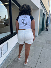 Load image into Gallery viewer, Full-body back view of a pair of size 22 Eloquii lightly distressed cream colored denim shorts styled with a white crop and white heels on a size 16/18 model. The photo is taken outside in natural lighting.
