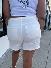 Load image into Gallery viewer, Back view of a pair of size 22 Eloquii lightly distressed cream colored denim shorts styled with a white crop and white heels on a size 16/18 model. The photo is taken outside in natural lighting.
