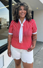Load image into Gallery viewer, Additional front view of a size 1/2X red and white vertical striped vintage blouse with white ascot scarf detail styled with cream shorts on a size 16/18 model. The photo is taken outside in natural lighting.
