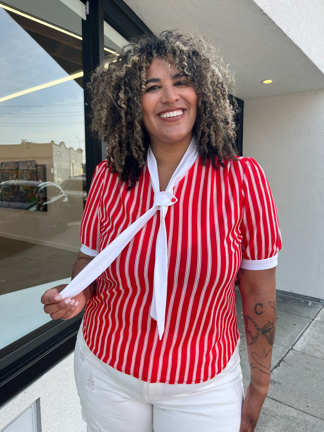Front view of a size 1/2X red and white vertical striped vintage blouse with white ascot scarf detail styled with cream shorts on a size 16/18 model. The photo is taken outside in natural lighting.