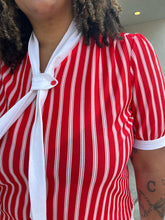 Load image into Gallery viewer, Close up view of the stripe pattern and ascot detail of a size 1/2X red and white vertical striped vintage blouse with white ascot scarf detail styled with cream shorts on a size 16/18 model. The photo is taken outside in natural lighting.
