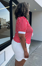 Load image into Gallery viewer, Side view of a size 1/2X red and white vertical striped vintage blouse with white ascot scarf detail styled with cream shorts on a size 16/18 model. The photo is taken outside in natural lighting.
