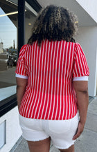 Load image into Gallery viewer, Back view of a size 1/2X red and white vertical striped vintage blouse with white ascot scarf detail styled with cream shorts on a size 16/18 model. The photo is taken outside in natural lighting.
