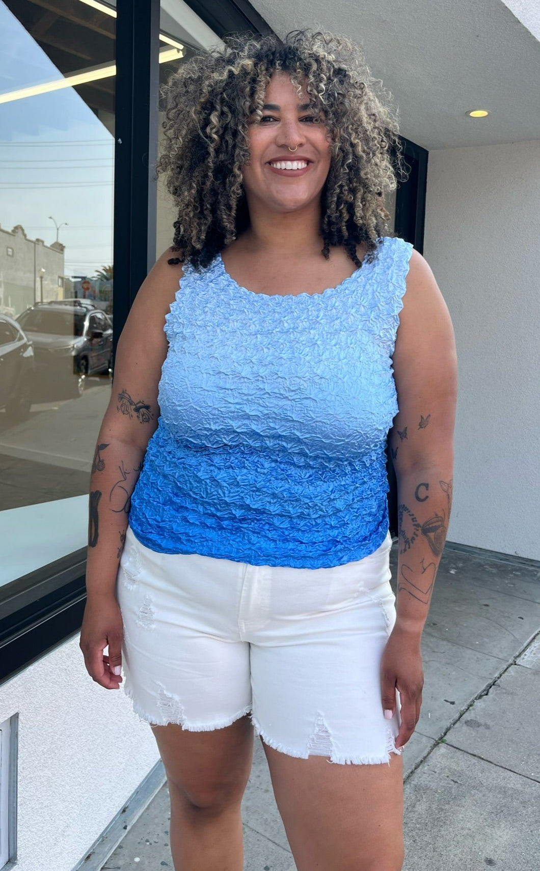 Front view of a size 1/2X light blue to dark blue ombre popcorn-style tank top styled over some cream denim shorts on a size 16/18 model. The photo is taken outside in natural lighting.