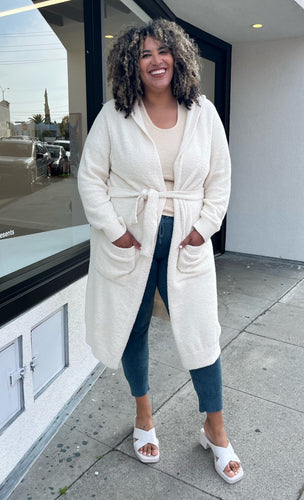 Full-body front view of a size 4 Fashion to Figure cozy cream-colored hooded fuzzy robe with pockets and tie belt styled over comfy clothes and white heels on a size 16/18 model. The photo is taken outside in natural lighting.