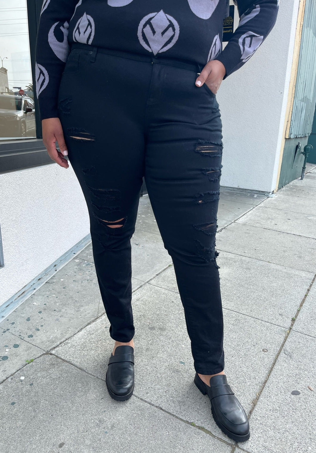Front view of a pair of size 18 Torrid black distressed skinny jeans styled with a black and gray sweater and black loafers on a size 16/18 model. The photo is taken outside in natural lighting.