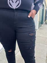 Load image into Gallery viewer, Close up view of the pocket and distressing of a pair of size 18 Torrid black distressed skinny jeans styled with a black and gray sweater and black loafers on a size 16/18 model. The photo is taken outside in natural lighting.
