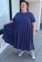 Load image into Gallery viewer, Additional full-body front view showing off the flounce of the skirt of a size 3/4X Anysize brand navy blue pleated baby doll t-shirt dress with pockets styled with white sneakers on a size 22/24 model. The photo is taken outside in natural lighting.
