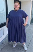 Load image into Gallery viewer, Final full-body front view of a size 3/4X Anysize brand navy blue pleated baby doll t-shirt dress with pockets styled with white sneakers on a size 22/24 model. The photo is taken outside in natural lighting.
