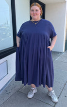 Load image into Gallery viewer, Full-body front view of a size 3/4X Anysize brand navy blue pleated baby doll t-shirt dress with pockets styled with white sneakers on a size 22/24 model. The photo is taken outside in natural lighting.
