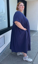 Load image into Gallery viewer, Full-body side view of a size 3/4X Anysize brand navy blue pleated baby doll t-shirt dress with pockets styled with white sneakers on a size 22/24 model. The photo is taken outside in natural lighting.
