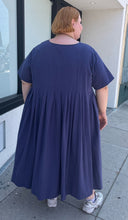 Load image into Gallery viewer, Full-body back view of a size 3/4X Anysize brand navy blue pleated baby doll t-shirt dress with pockets styled with white sneakers on a size 22/24 model. The photo is taken outside in natural lighting.
