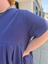 Load image into Gallery viewer, Close up view of the breathable cotton fabric of a size 3/4X Anysize brand navy blue pleated baby doll t-shirt dress with pockets on a size 22/24 model. The photo is taken outside in natural lighting.
