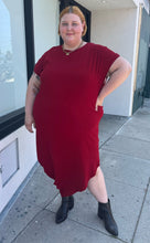 Load image into Gallery viewer, Full-body front view of a size 24 Universal Standard deep red asymmetrical t-shirt maxi dress with pockets styled with black boots on a size 22/24 model. The photo is taken outside in natural lighting.
