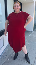 Load image into Gallery viewer, Additional full-body front view of a size 24 Universal Standard deep red asymmetrical t-shirt maxi dress with pockets styled with black boots on a size 22/24 model. The photo is taken outside in natural lighting.
