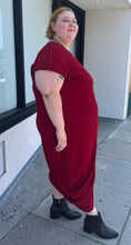 Load image into Gallery viewer, Full-body side view of a size 24 Universal Standard deep red asymmetrical t-shirt maxi dress with pockets styled with black boots on a size 22/24 model. The photo is taken outside in natural lighting.
