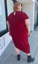 Load image into Gallery viewer, Full-body back view of a size 24 Universal Standard deep red asymmetrical t-shirt maxi dress with pockets styled with black boots on a size 22/24 model. The photo is taken outside in natural lighting.
