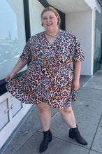 Load image into Gallery viewer, Additional full-body front view of a size 28 (fits like 24/26) Simply Be pink, blue, orange, white, and black leopard pattern faux wrap mini dress with a tulip hem styled with black boots on a size 22/24 model. The photo is taken outside in natural lighting.
