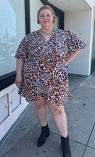 Load image into Gallery viewer, Full-body front view of a size 28 (fits like 24/26) Simply Be pink, blue, orange, white, and black leopard pattern faux wrap mini dress with a tulip hem styled with black boots on a size 22/24 model. The photo is taken outside in natural lighting.
