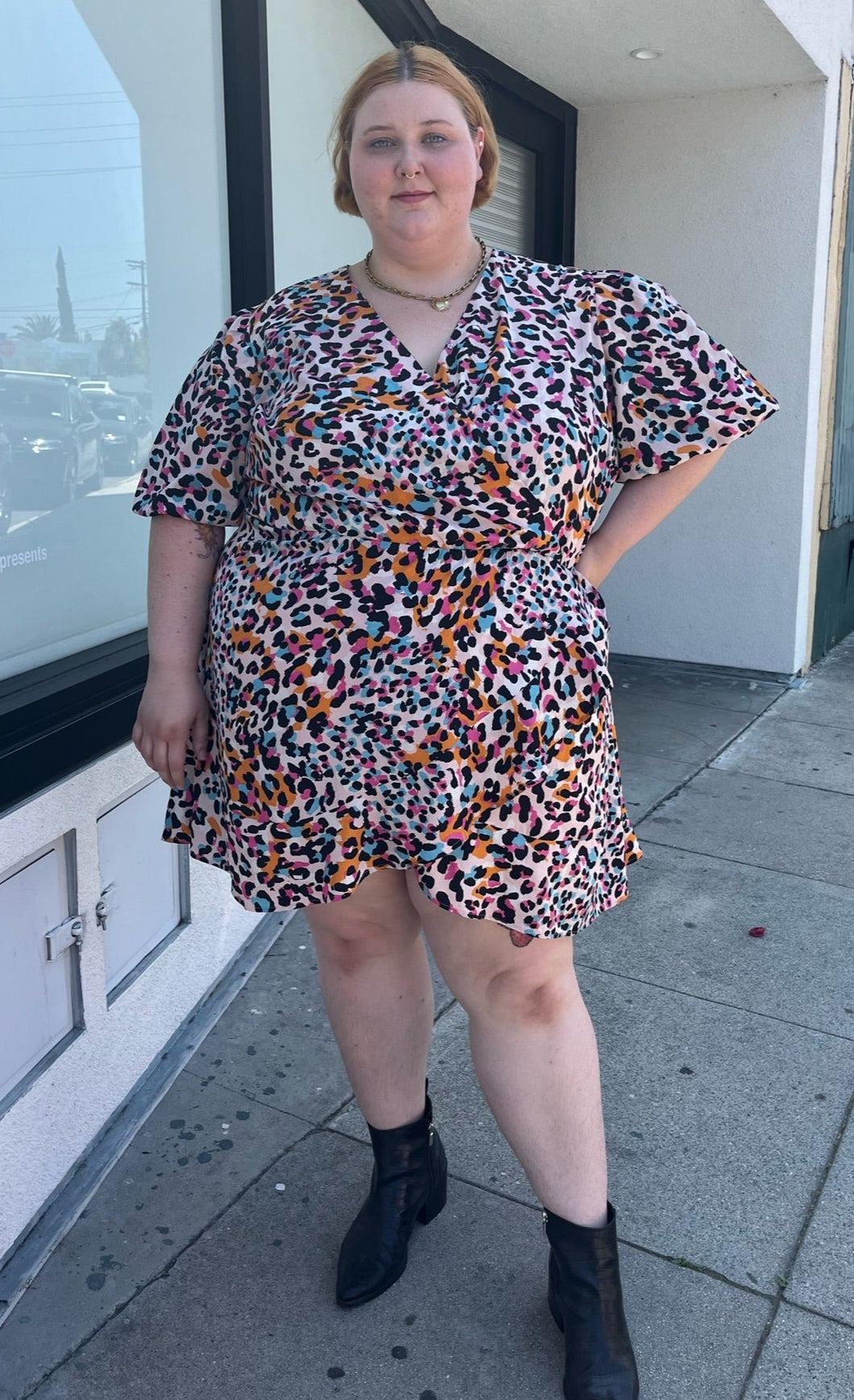 Full-body front view of a size 28 (fits like 24/26) Simply Be pink, blue, orange, white, and black leopard pattern faux wrap mini dress with a tulip hem styled with black boots on a size 22/24 model. The photo is taken outside in natural lighting.
