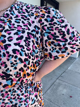 Load image into Gallery viewer, Close up view of the pale pink background and pink, orange, and blue multicolor leopard pattern of a size 28 (fits like 24/26) Simply Be pink, blue, orange, white, and black leopard pattern faux wrap mini dress with a tulip hem on a size 22/24 model. The photo is taken outside in natural lighting.
