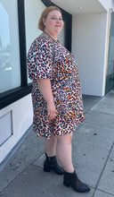Load image into Gallery viewer, Full-body side view of a size 28 (fits like 24/26) Simply Be pink, blue, orange, white, and black leopard pattern faux wrap mini dress with a tulip hem styled with black boots on a size 22/24 model. The photo is taken outside in natural lighting.
